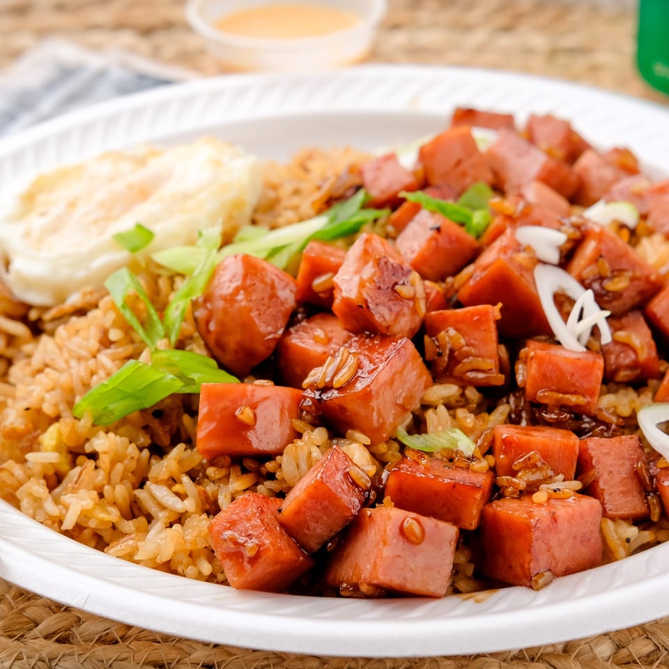 Plate of fried rice topped with diced sausage, vegetables, creamy sauce, and green onions, served with chopsticks.