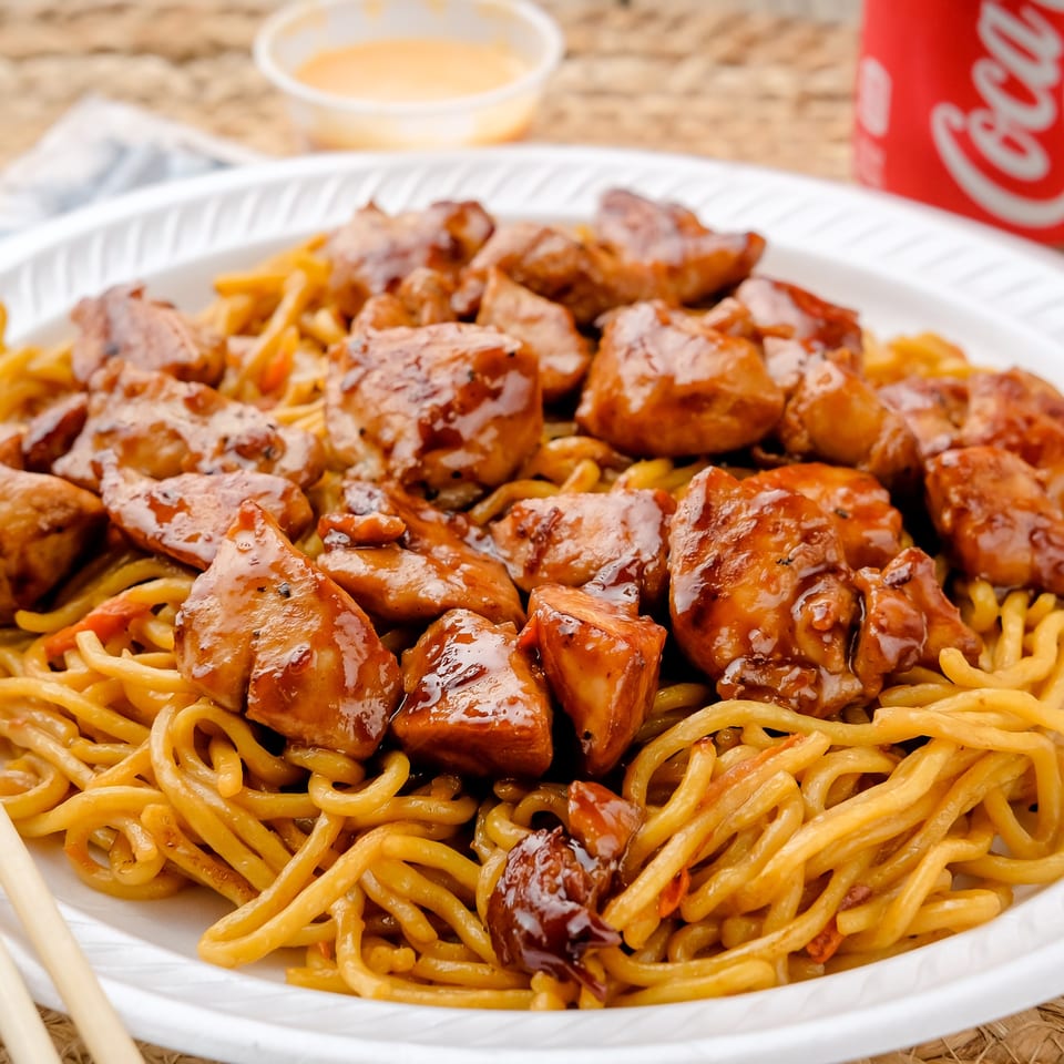 Glazed chicken pieces served over stir-fried noodles with chopsticks, dipping sauce, and a Coca-Cola cup.