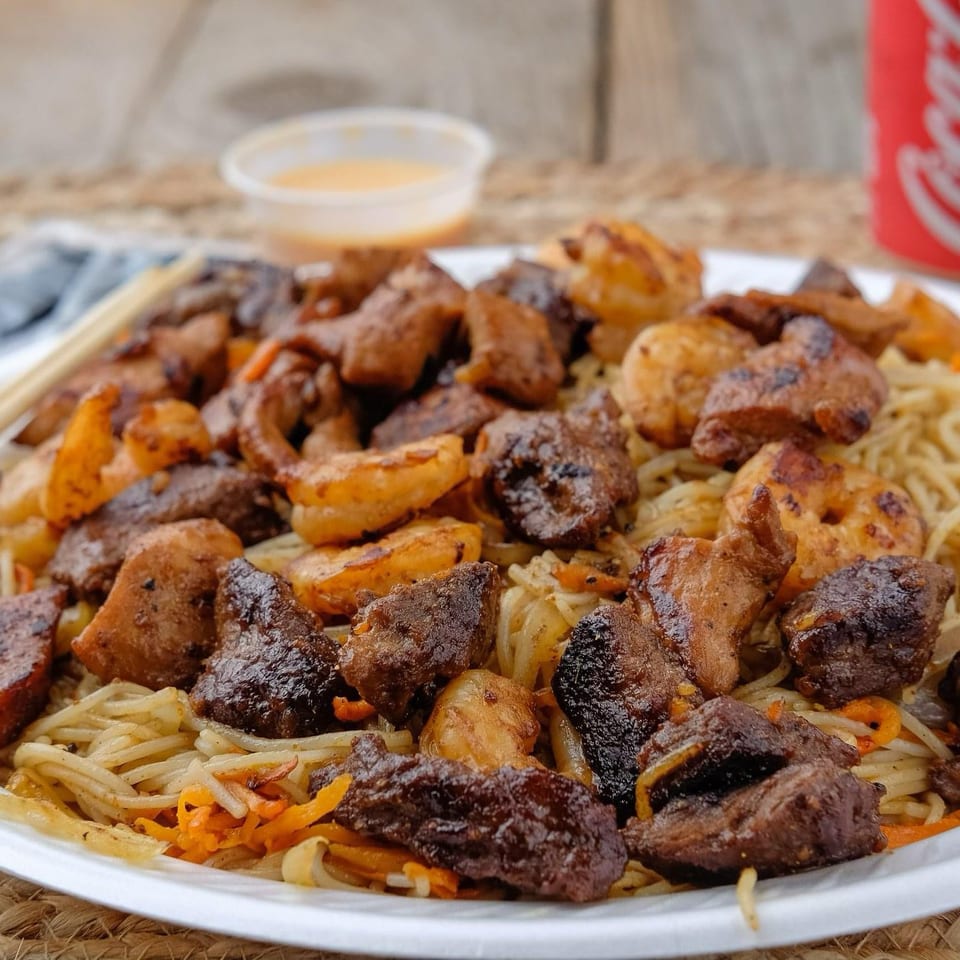 Plate of grilled meats and shrimp over noodles with dipping sauce, chopsticks, and a Coca-Cola can.