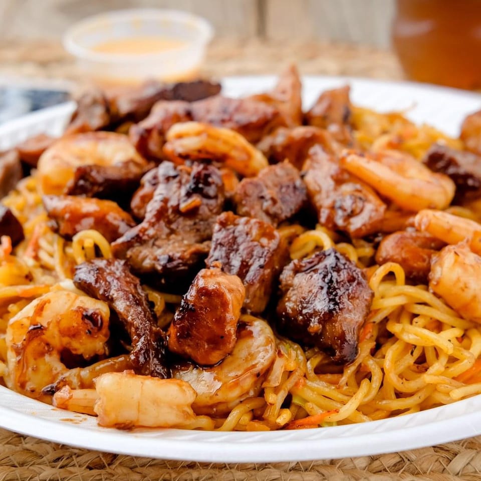 Plate of saucy noodles with shrimp, grilled meat, vegetables, chopsticks, dipping sauce, and a beverage.